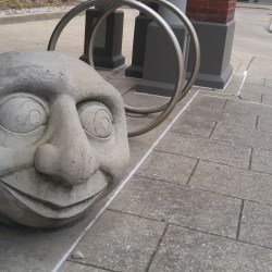 Urban bike rack art installation. A monumental concrete head on the ground looking out the corner of his eye, with two bike rings behind, making him look a bit like a modern art caterpillar.