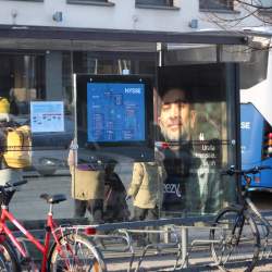 Back of person in a glass bus stop with screen and reflections of screens and in back of bus stop/foreground, bicycles, and on right back of bus