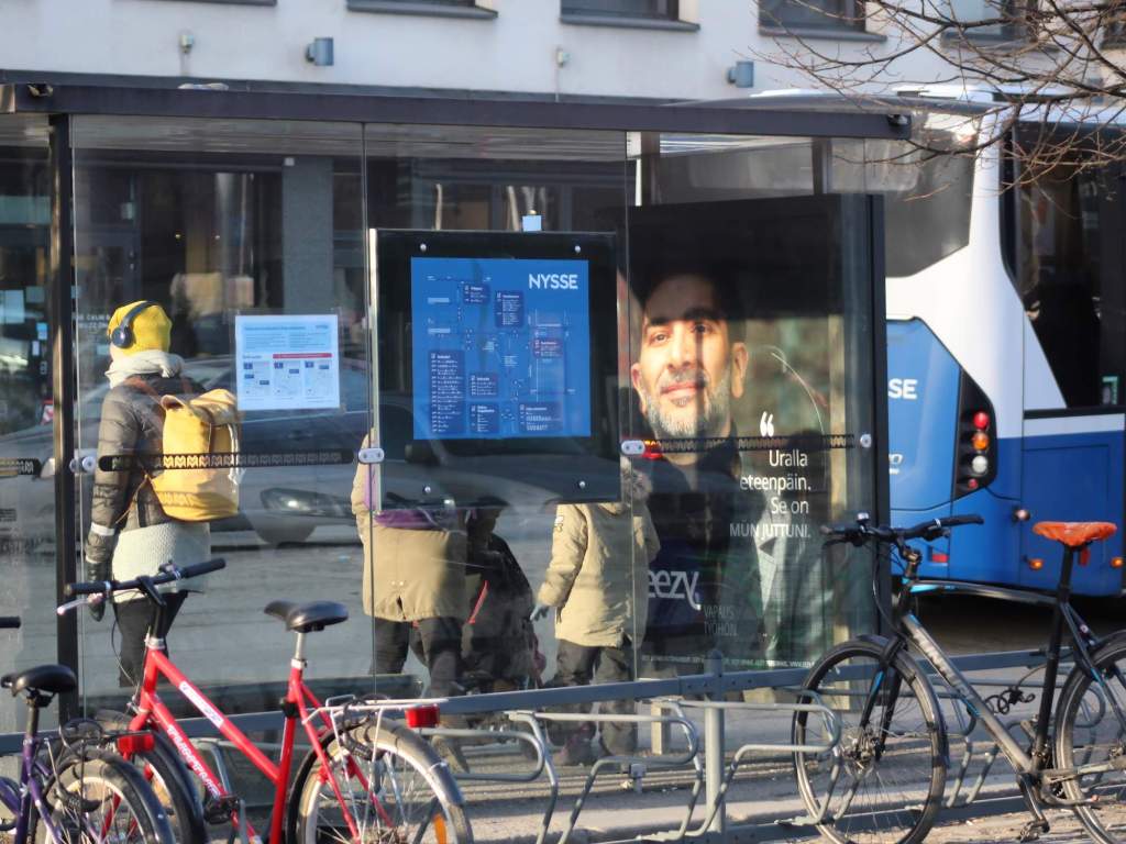 Back of person in a glass bus stop with screen and reflections of screens and in back of bus stop/foreground, bicycles, and on right back of bus