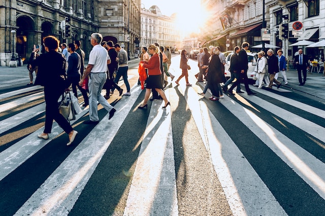 city street people walking crosswalk, bright sun between buildings